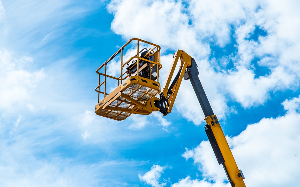 Hydraulic lift platform with bucket of yellow construction vehicle, heavy industry, blue sky and white clouds on background 1050x656