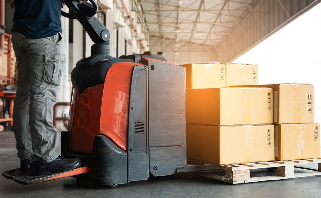 Shipment boxes, Cargo warehousing. Worker driving electric forklift pallet jack unloading cardboard boxes on pallet 1050x656