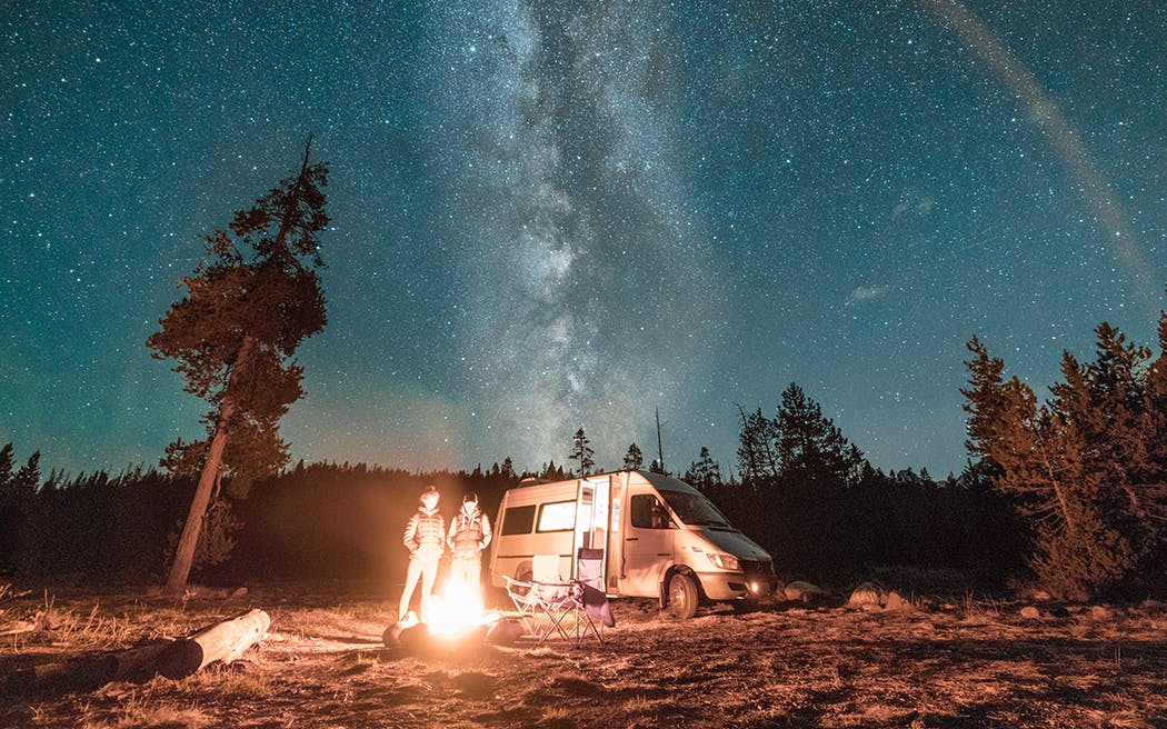 Vanlife couple standing in front of campfire and van under Milky Way night sky 1050x656